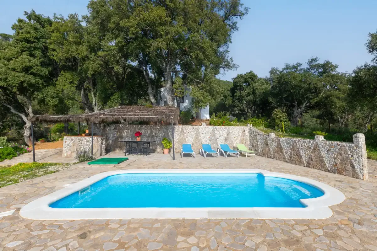 An outdoor swimming pool sits beside a stone patio and shaded seating area.