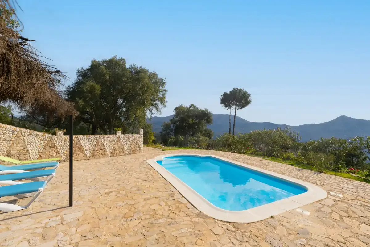 An outdoor swimming pool sits on a stone-paved terrace with hills in the distance.