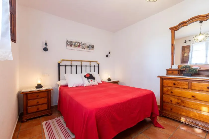 A bedroom with a red bedspread, bedside tables, and a wooden dresser with mirror.