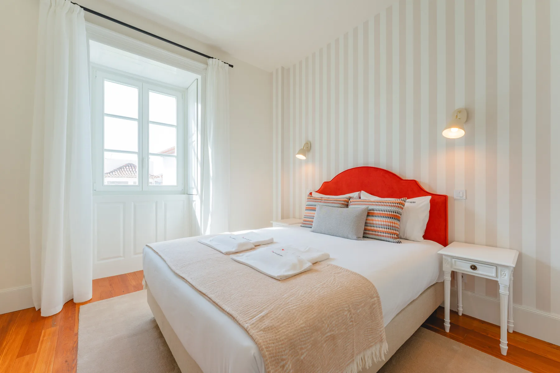 A bright bedroom with a bed, striped wallpaper, and a large window.