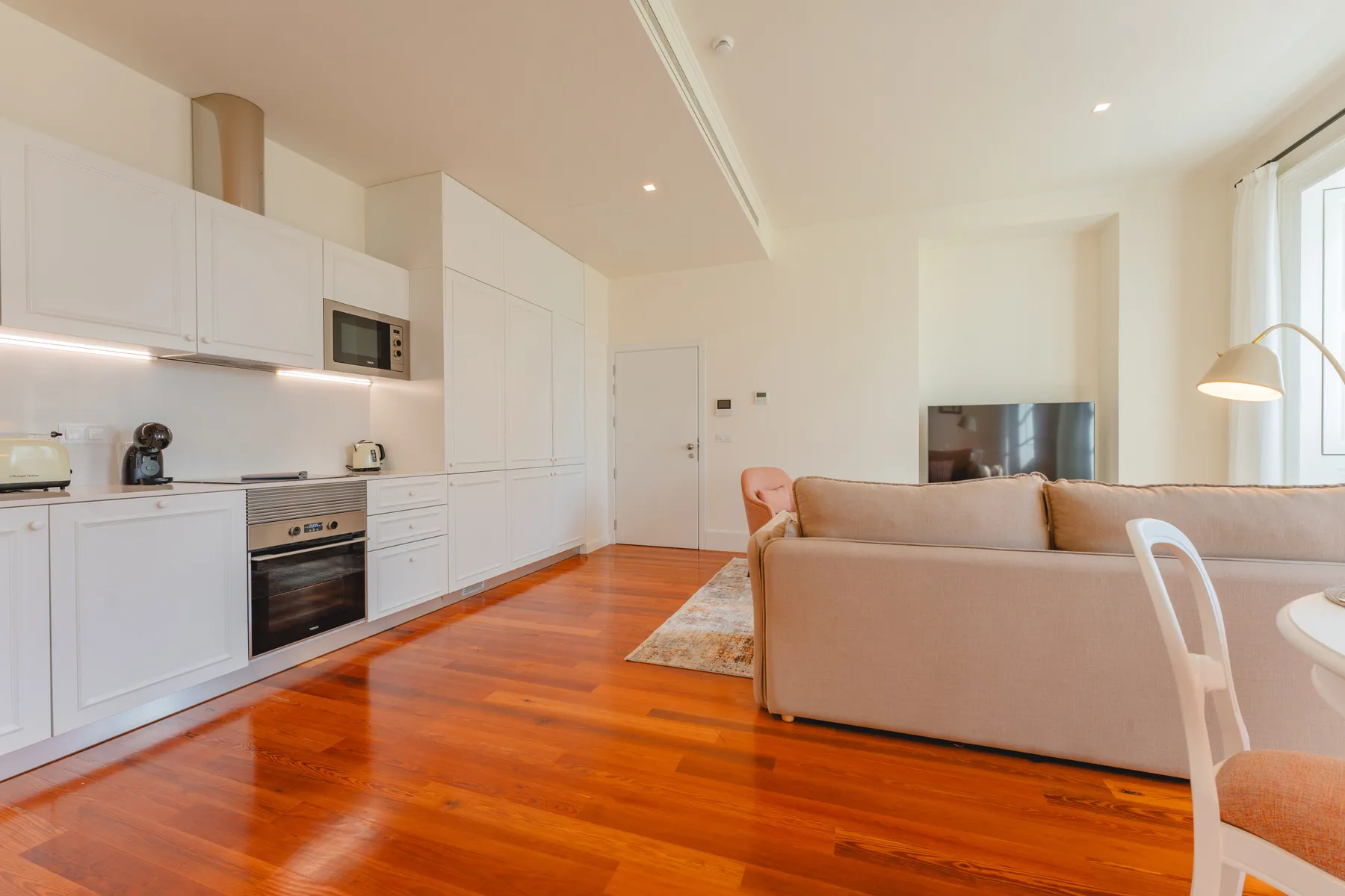 An open-plan living room and kitchen with white cabinets and warm wood flooring.