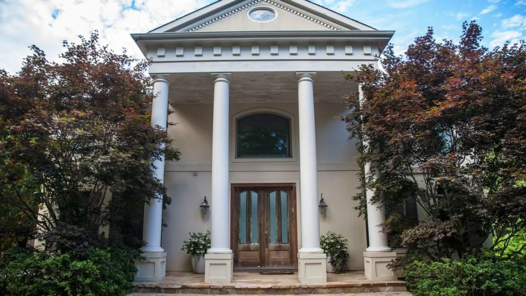 Entrada de un edificio con columnas y puertas dobles de madera, enmarcada por árboles.