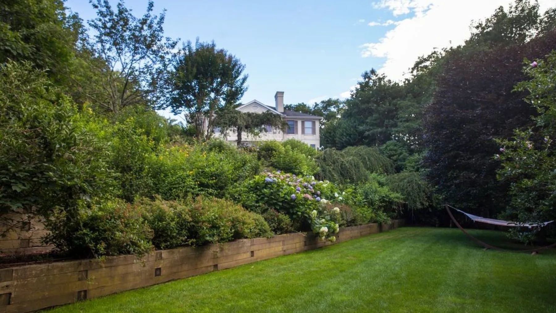 Un jardín con césped verde, arbustos floridos y una casa al fondo.