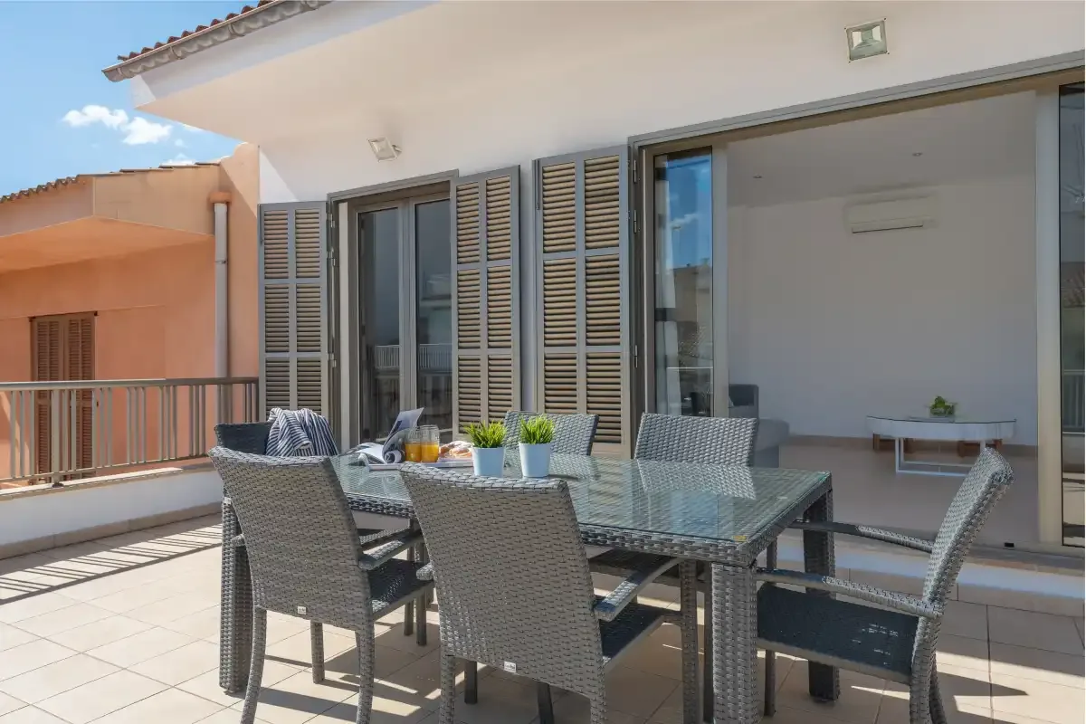 A sunny terrace with a glass dining table and seating beside open doors.