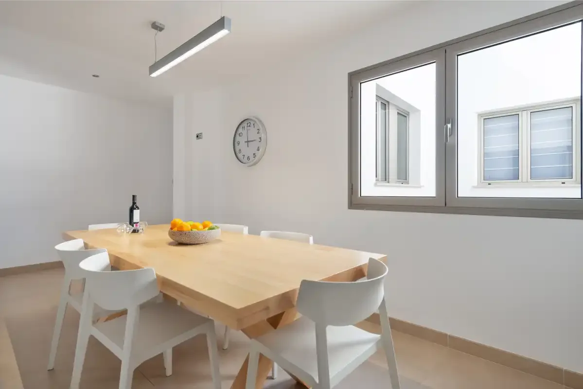 A bright dining area has a wooden table, white seating, and large windows.