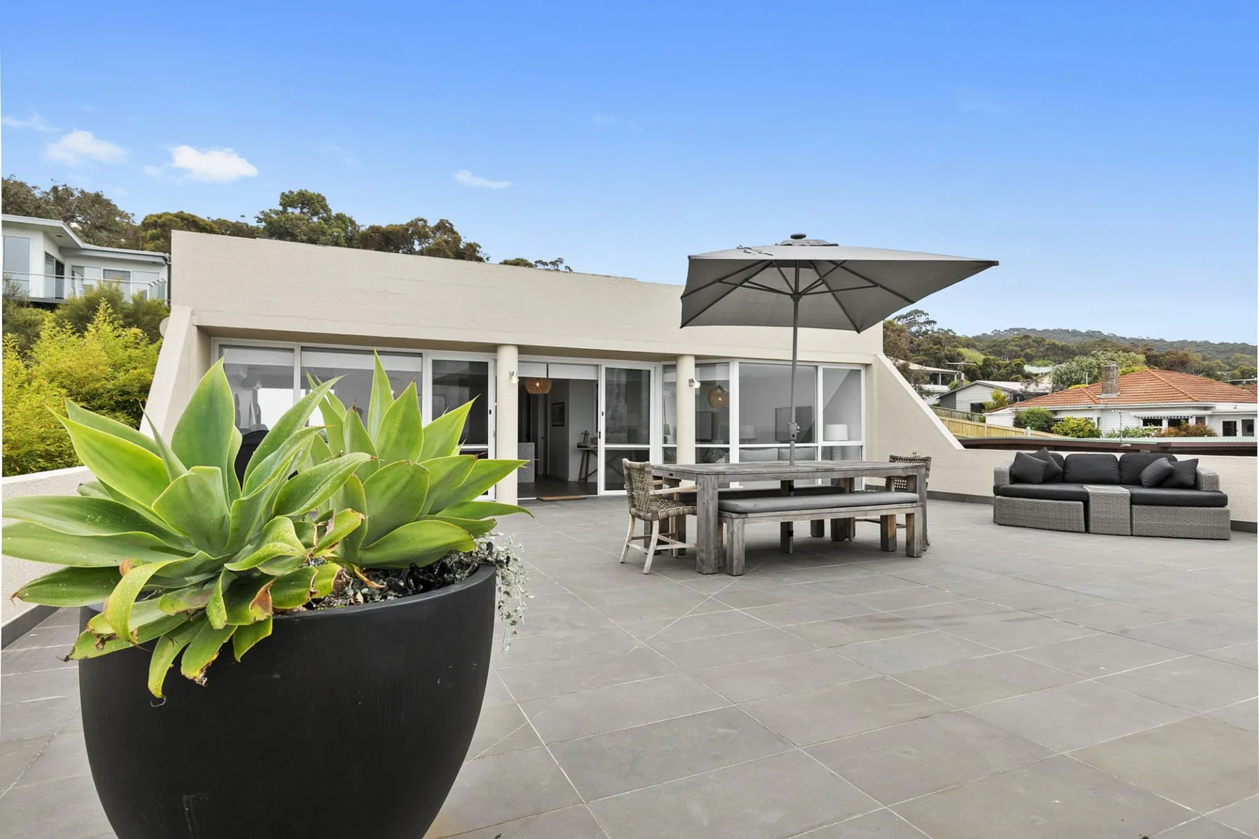 Un amplio patio exterior tiene una mesa de comedor, asientos y una gran planta en maceta.