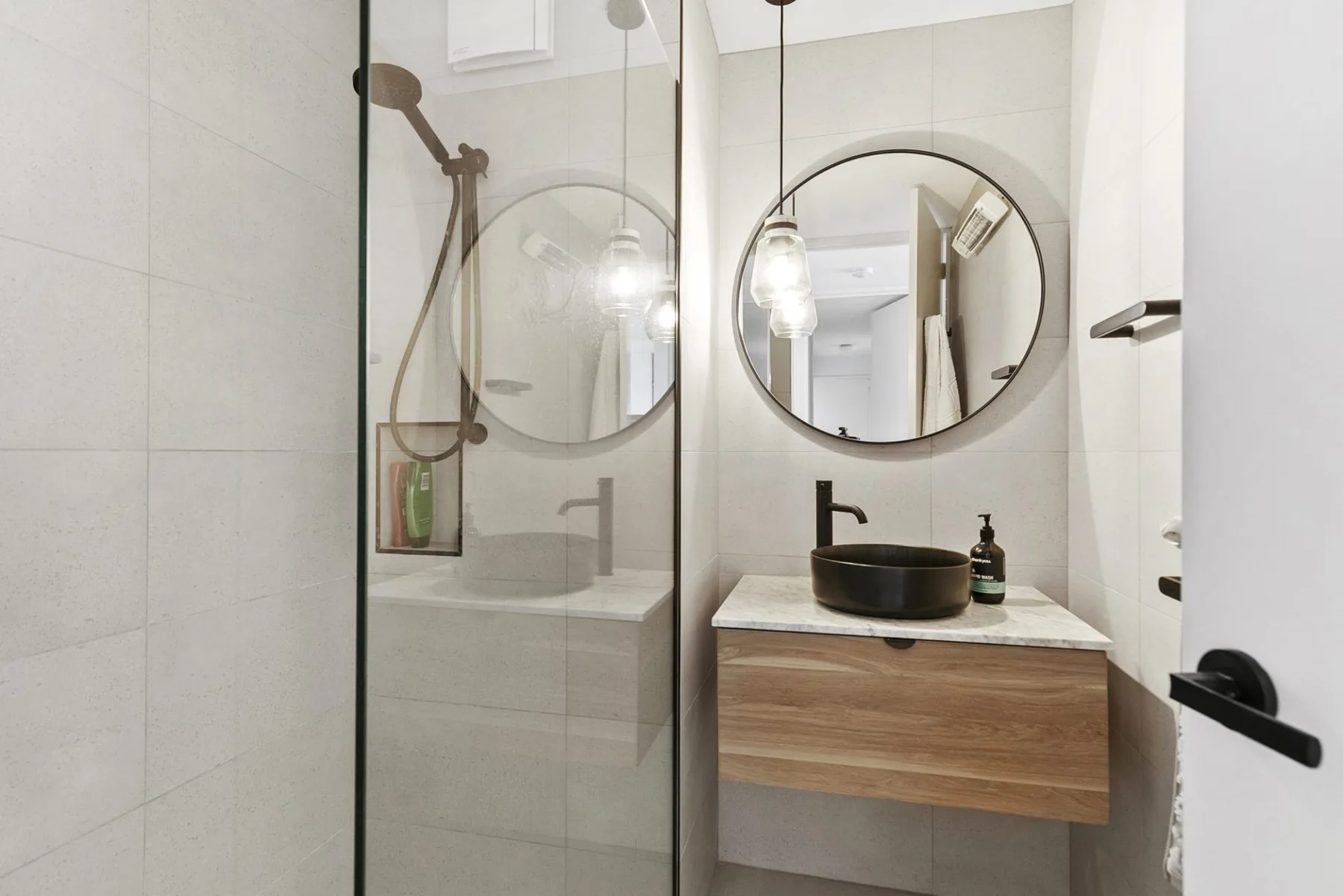 Un baño luminoso con una ducha de vidrio y un lavabo bajo un espejo redondo.