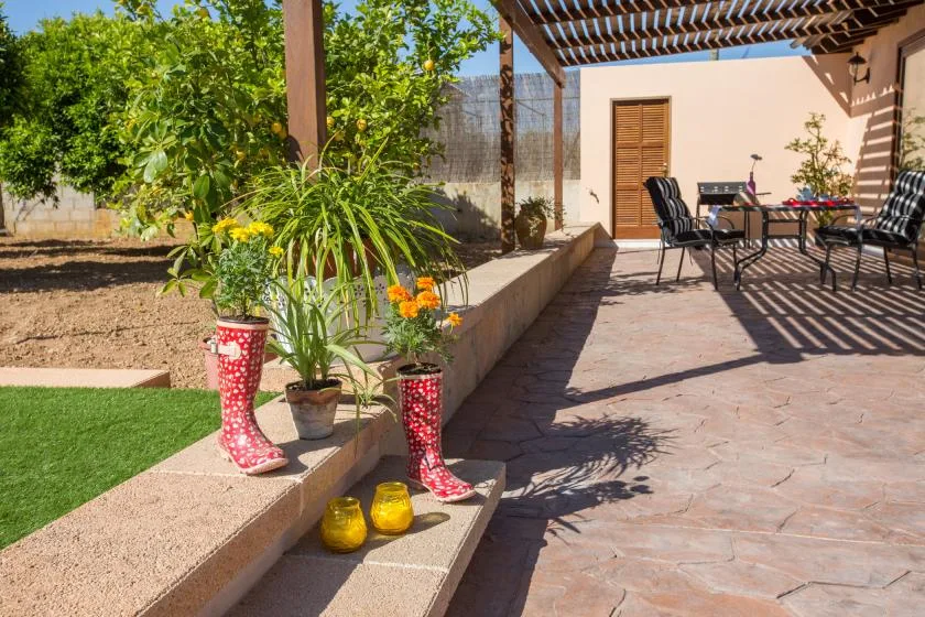 A paved patio with outdoor seating sits beside a planted garden under a pergola.