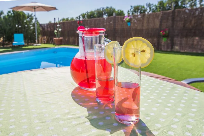 A table with a pitcher and glasses of red drink sits beside a swimming pool.