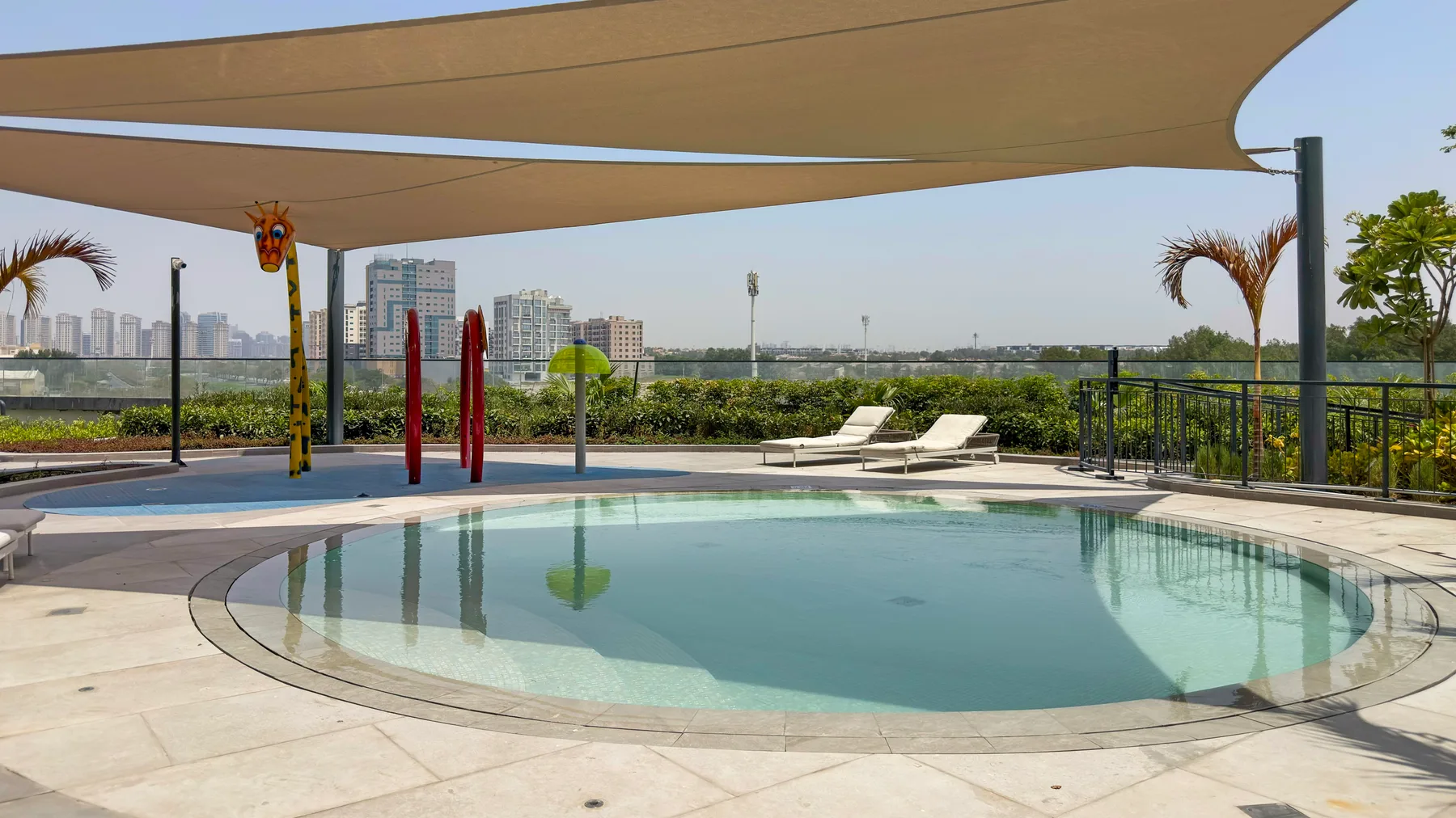 Una piscina al aire libre está bajo una cubierta de sombra, con asientos y el horizonte urbano al fondo.