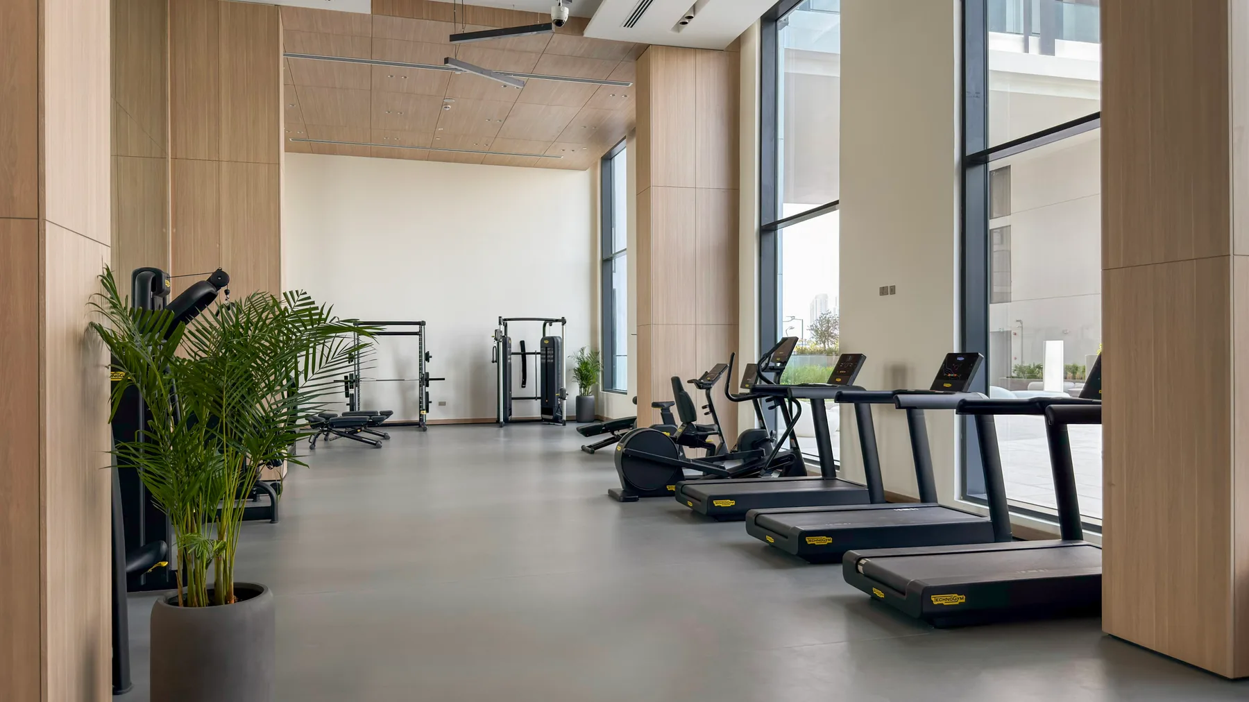 Un gimnasio luminoso con cintas de correr y equipos de ejercicio junto a grandes ventanales.