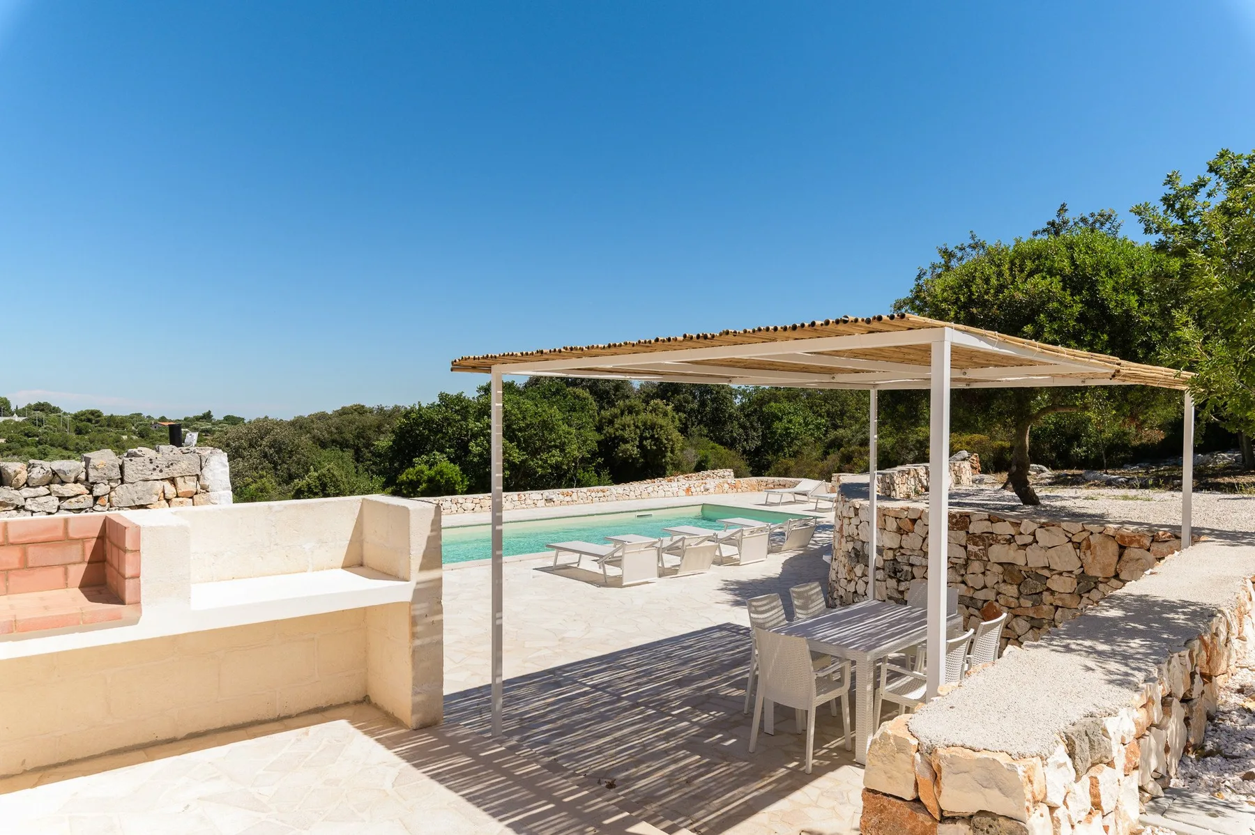 Zona de piscina al aire libre con un comedor a la sombra junto a un muro de piedra.