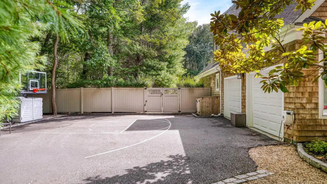 Una cancha de baloncesto al aire libre junto a una casa, rodeada de árboles.