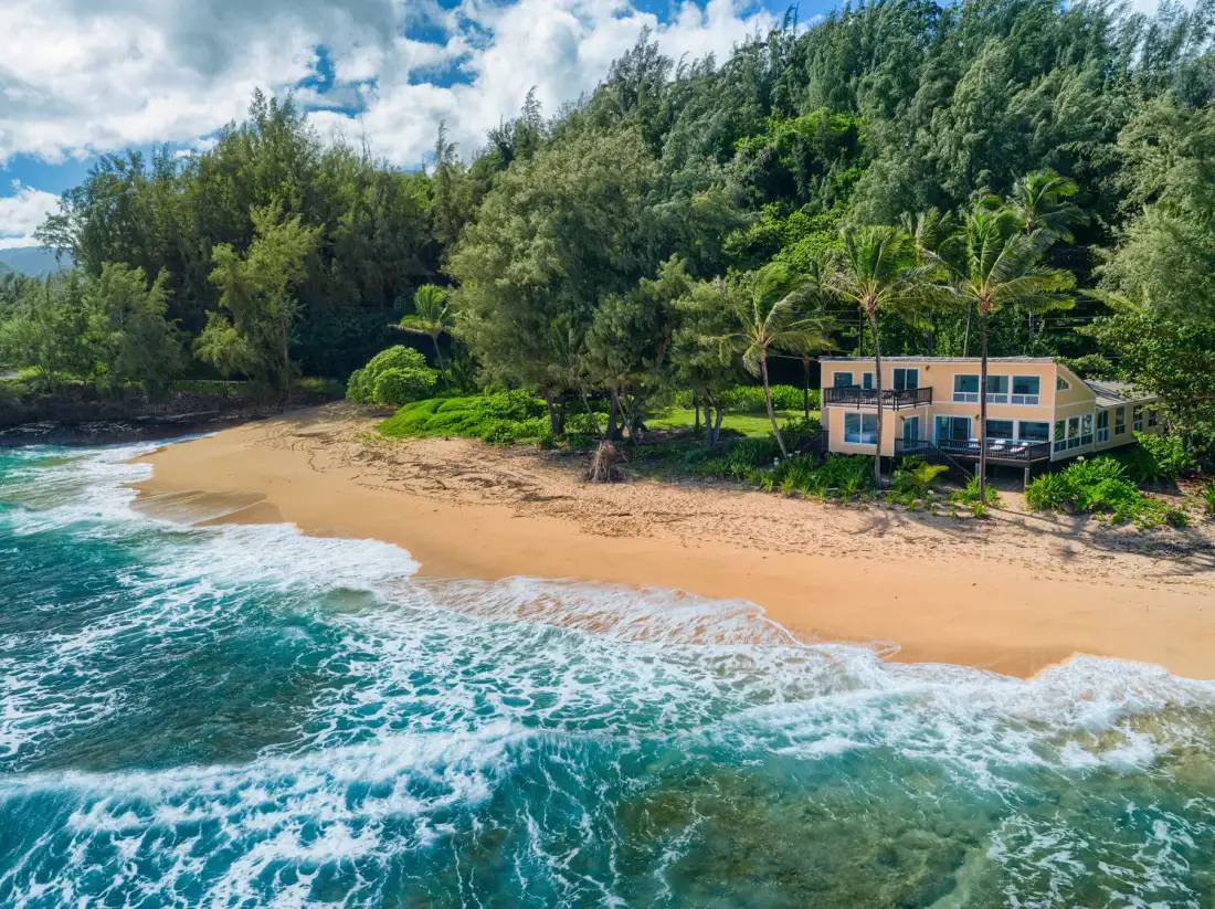 Un edificio junto a la playa se encuentra al lado de una orilla arenosa, con olas y vegetación densa.