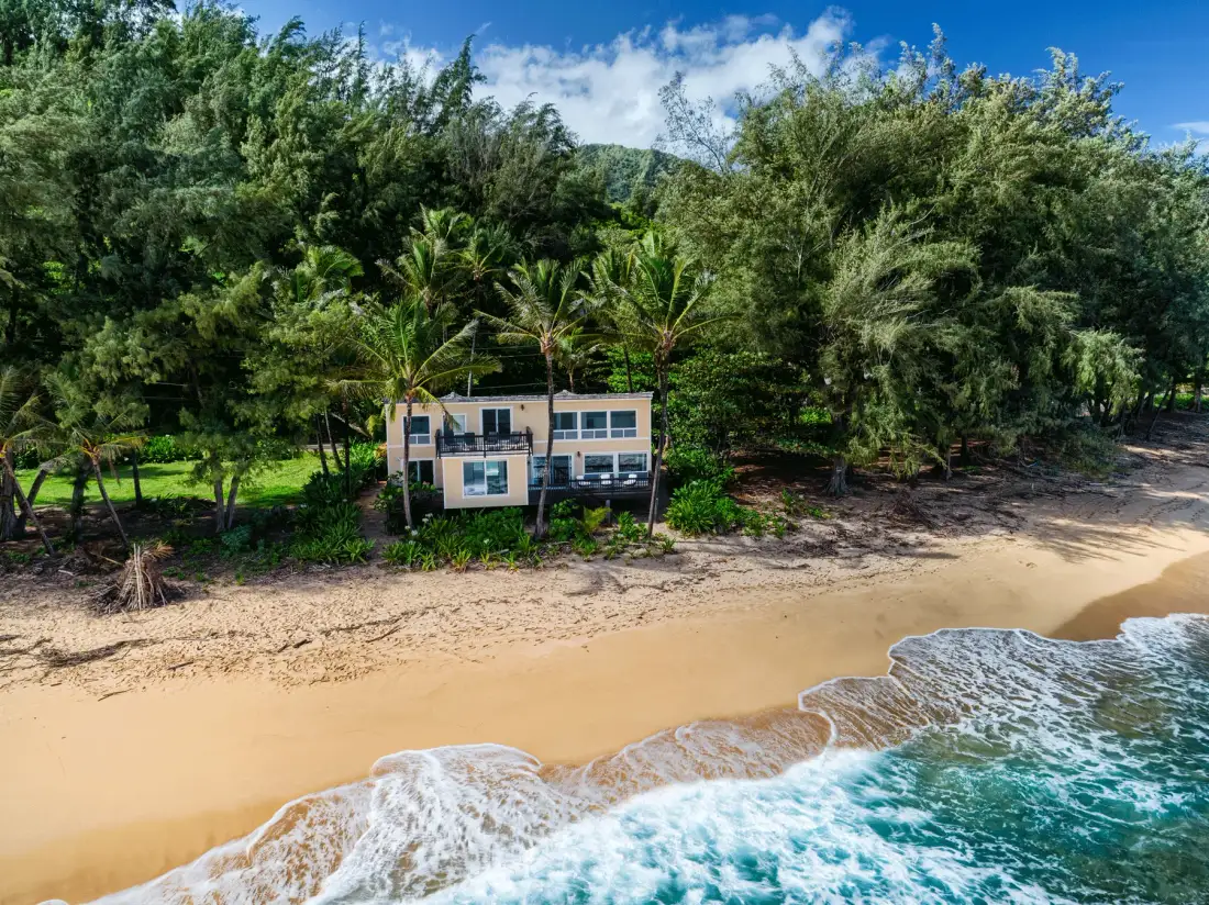 Un edificio junto a la playa está rodeado de árboles, arena y olas.