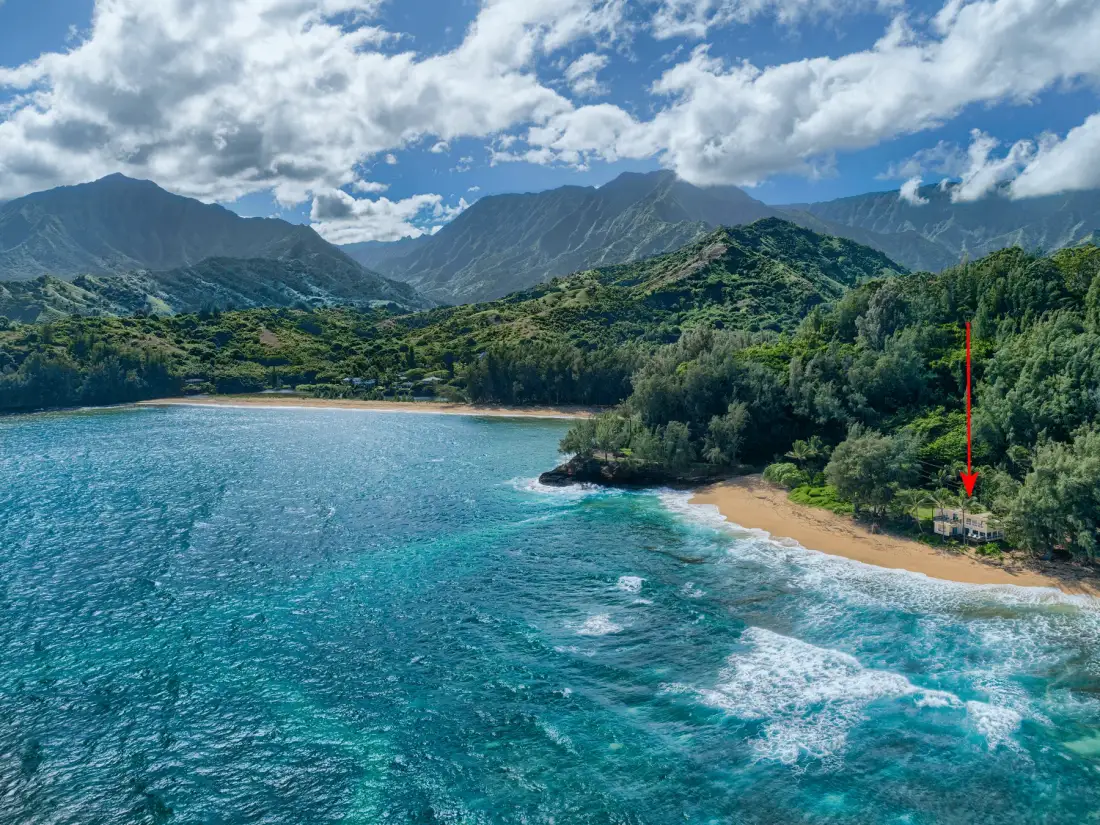 Una costa turquesa se encuentra con una playa de arena bajo montañas verdes y un cielo nublado.