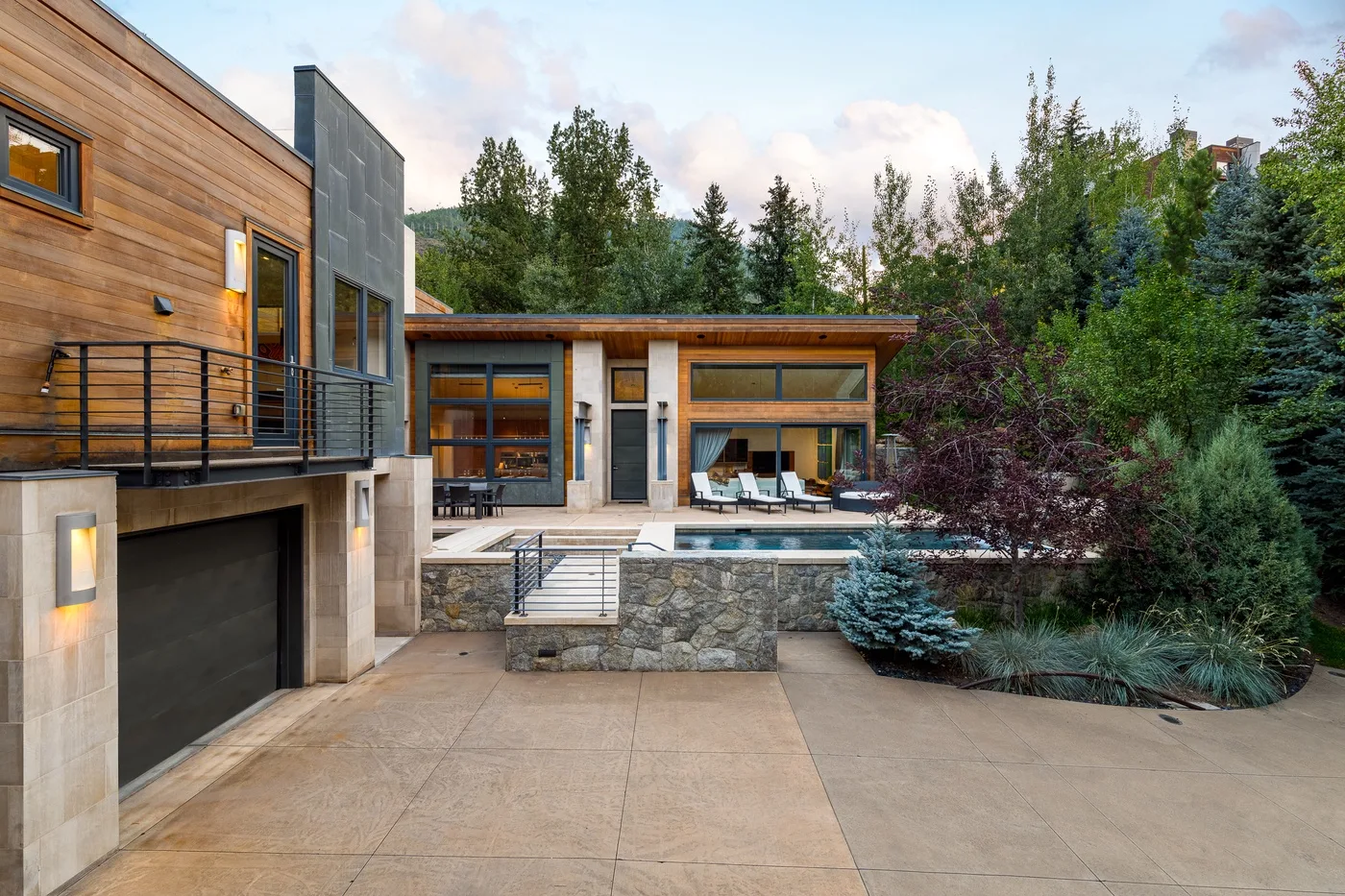 A modern house opens onto a paved courtyard, with trees and landscaping.