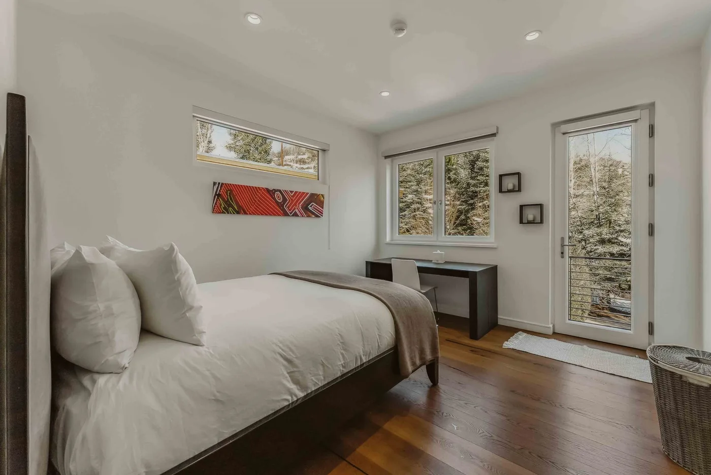 A bright bedroom with a bed, desk, and windows overlooking trees.