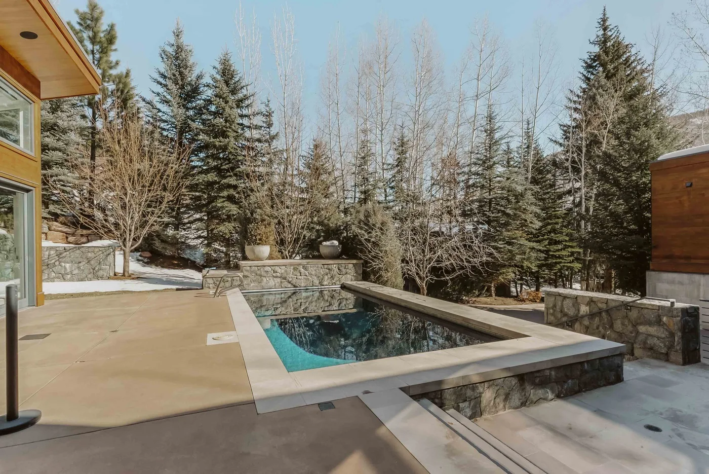 An outdoor swimming pool sits beside a paved patio, with trees in the background.