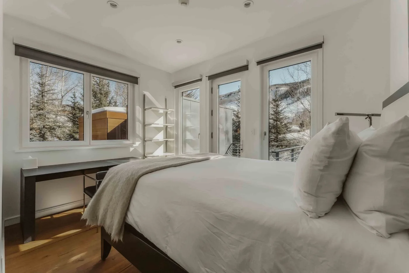 A bright bedroom with a bed beside large windows overlooking trees and mountains.