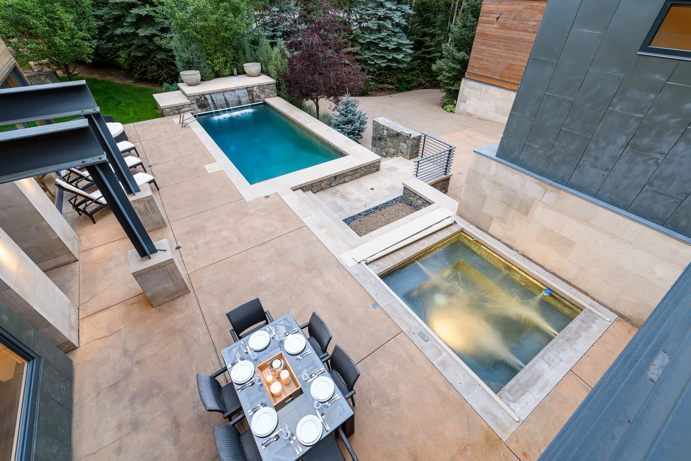 An outdoor patio has a dining table beside a swimming pool and a smaller water feature.
