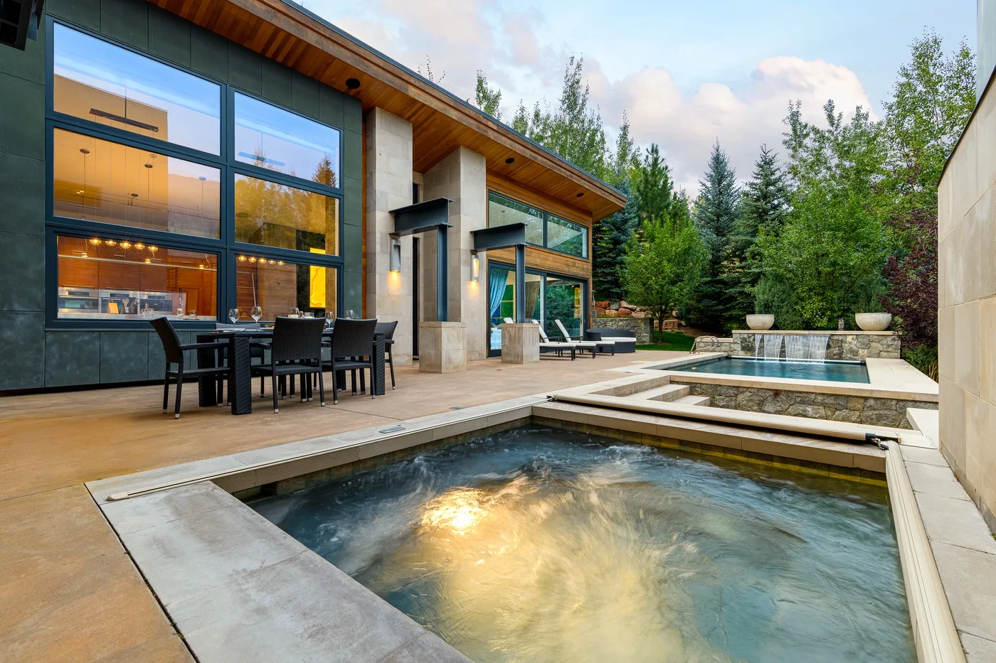 A modern house opens onto a patio with a table and outdoor pools.