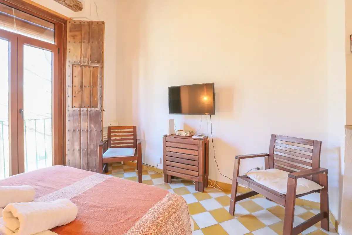 A bedroom with a bed, two wooden seats, and a television on the wall.