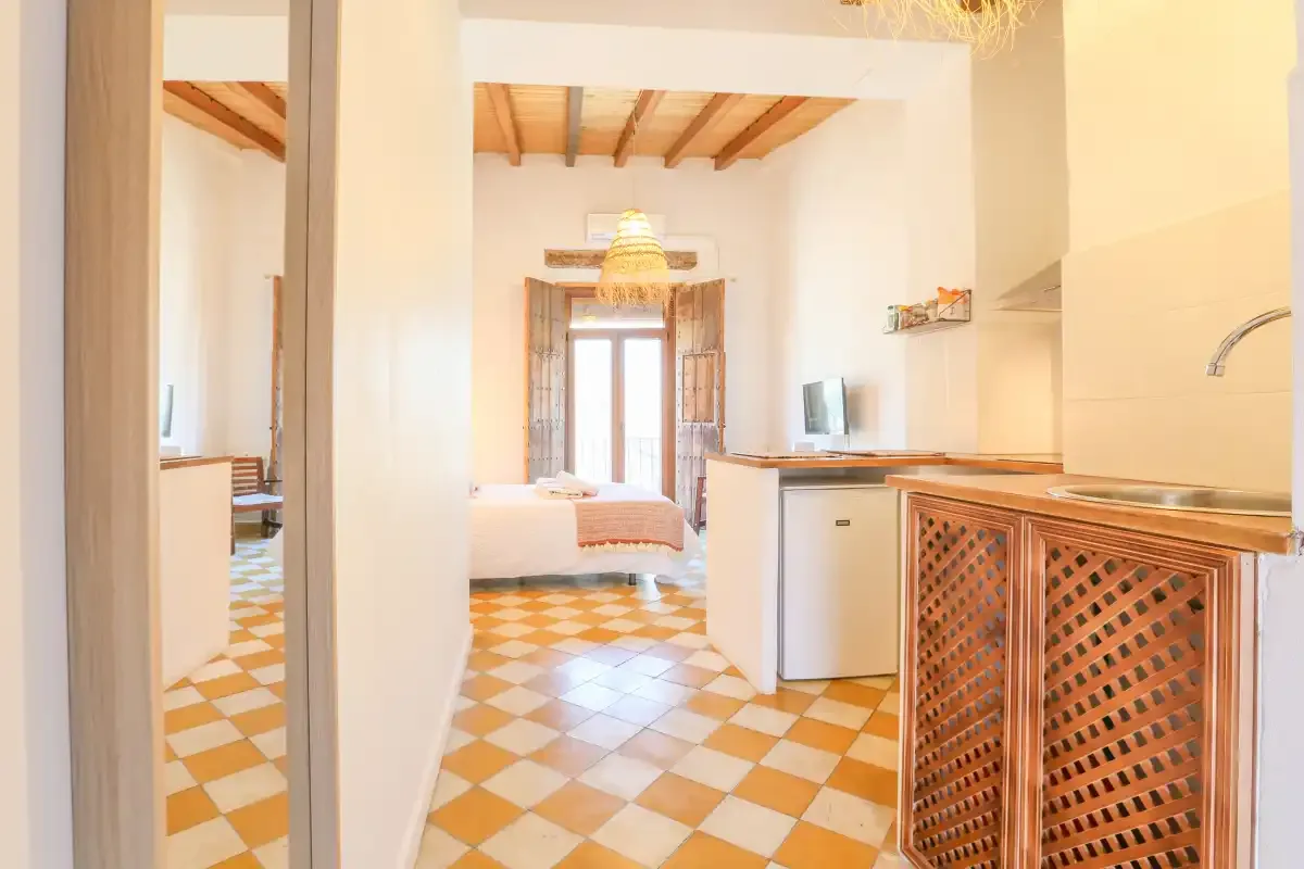 A bright open-plan room combines a sleeping area, kitchen counter, and patterned tile floor.