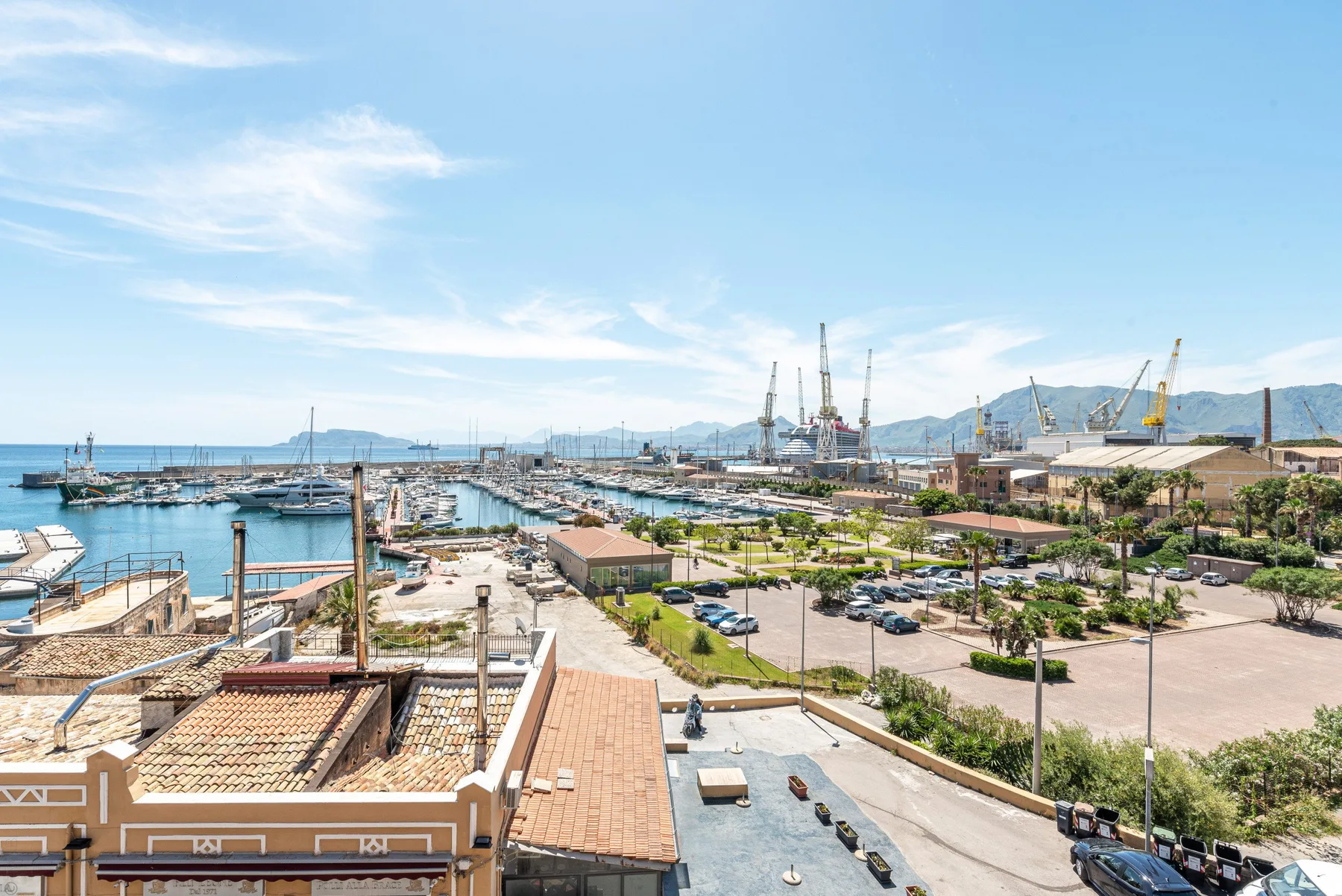 Un puerto deportivo y paseo marítimo con barcos, senderos ajardinados y grúas al fondo.