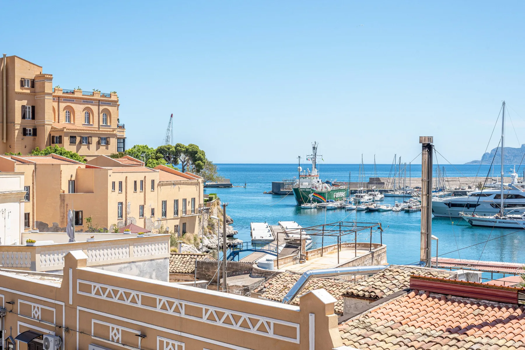 Un puerto costero soleado con barcos amarrados junto a edificios frente al mar.