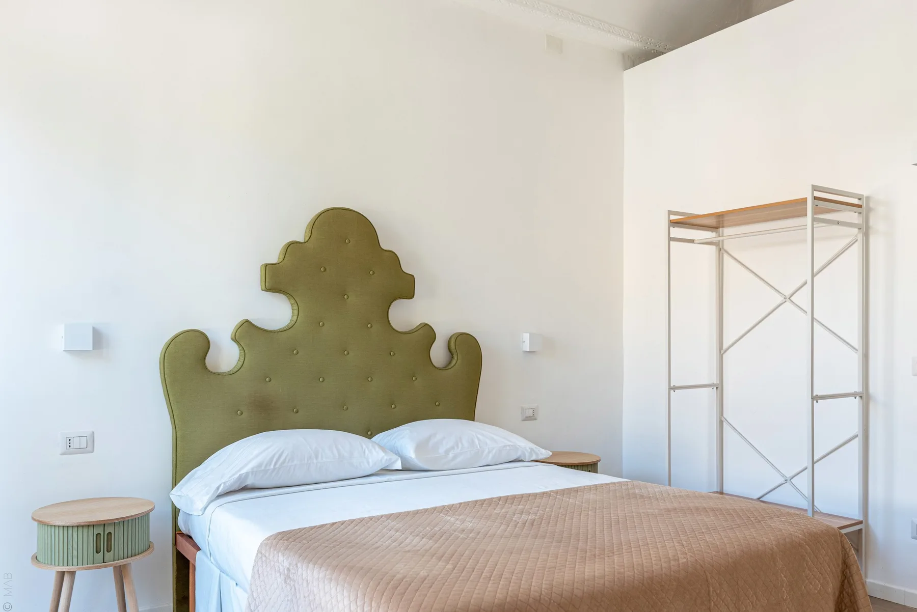 Un dormitorio con una cama, cabecero tapizado verde oliva y estantería metálica abierta.