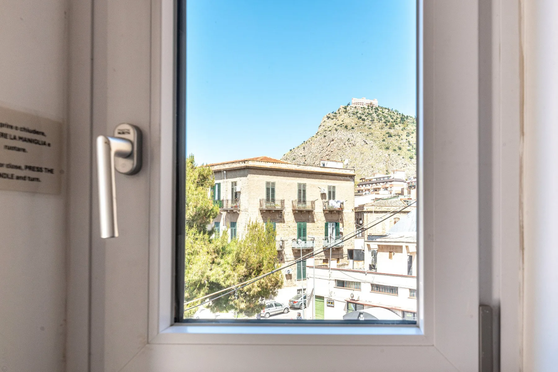 Una ventana da a edificios cercanos y una colina rocosa bajo un cielo despejado.