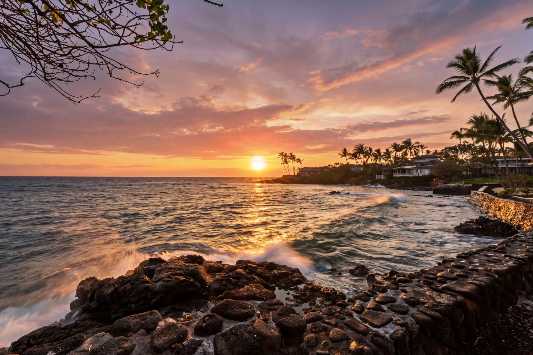 Una costa rocosa y palmeras enmarcan la puesta de sol sobre el océano.