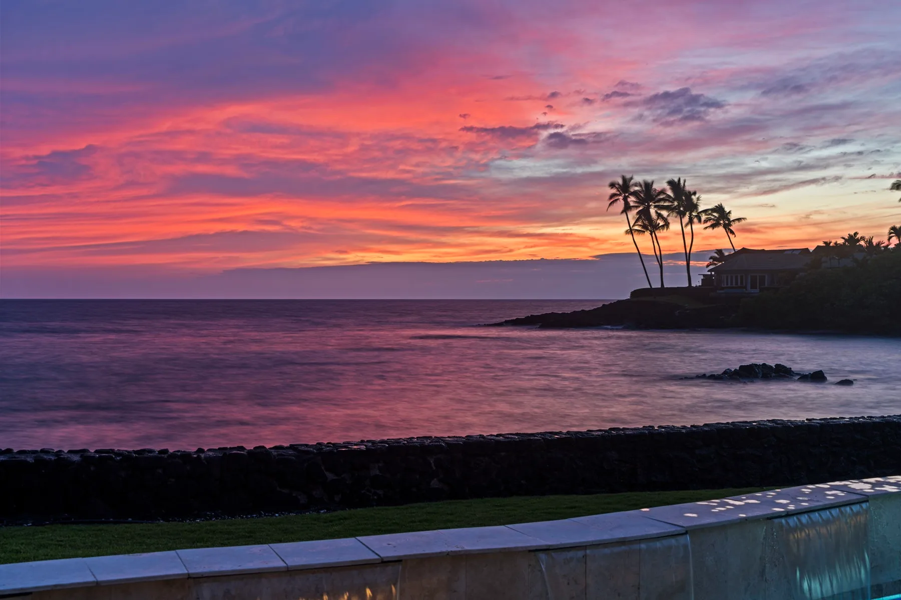 Un atardecer costero tiñe el cielo sobre el océano, con palmeras junto a la orilla.