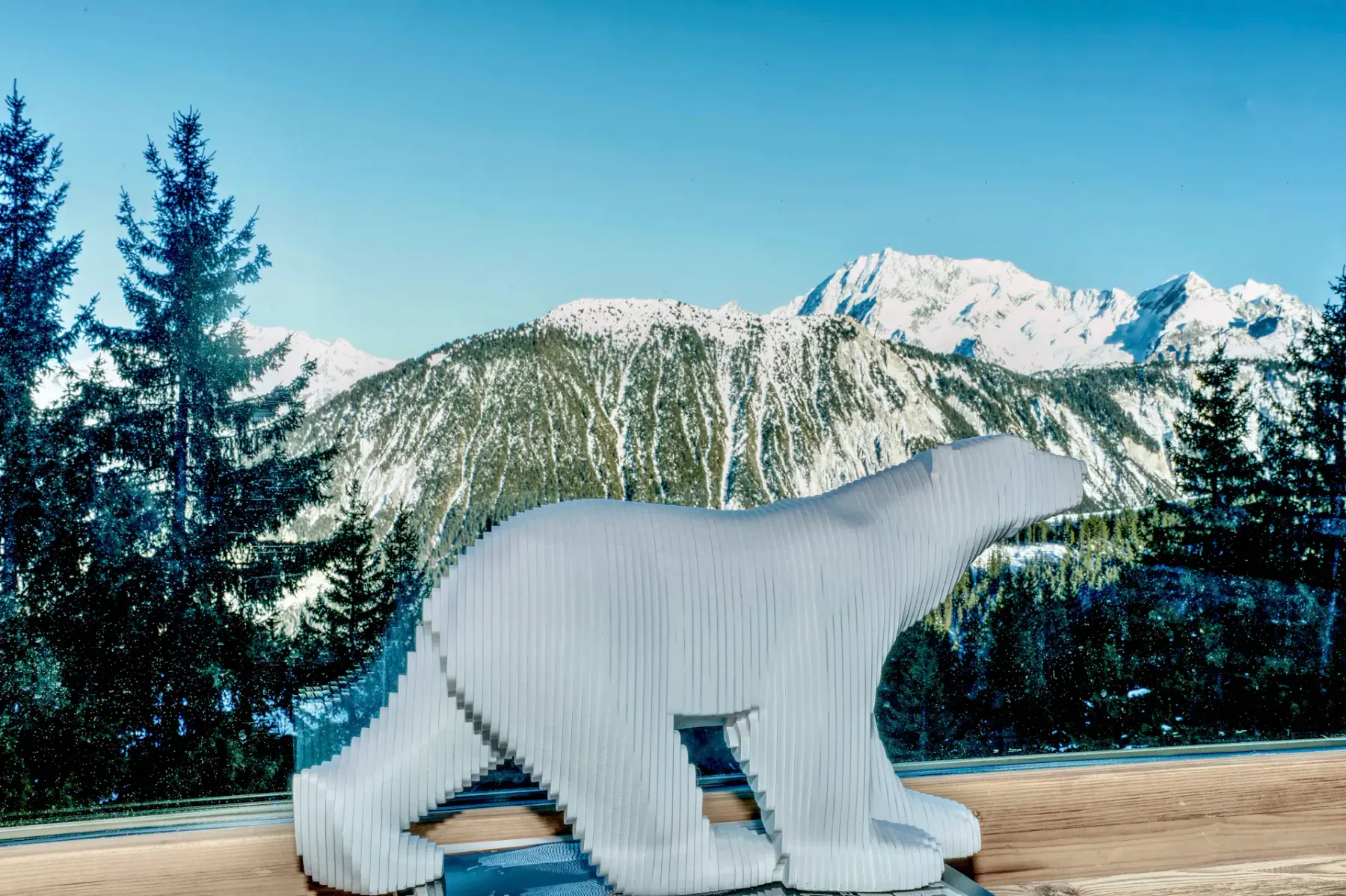 Una escultura blanca de un oso polar sobre el alféizar de una ventana, con un paisaje de montañas nevadas y pinos al fondo.