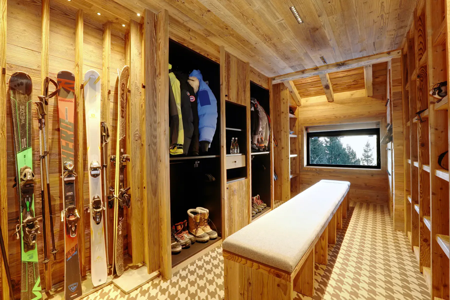 Cálida sala para guardar material de esquí, revestida de madera, con soportes para esquís, tablas de snowboard, botas y equipo. Un largo banco acolchado recorre el centro. Gran ventana con vistas.
