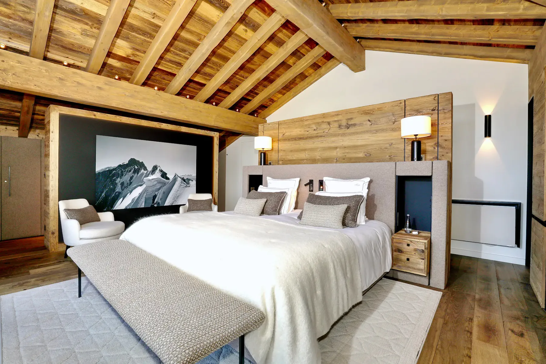 Acogedor dormitorio con vigas de madera, cama grande, lámparas modernas, una fotografía de montaña en la pared y un banco a los pies de la cama.