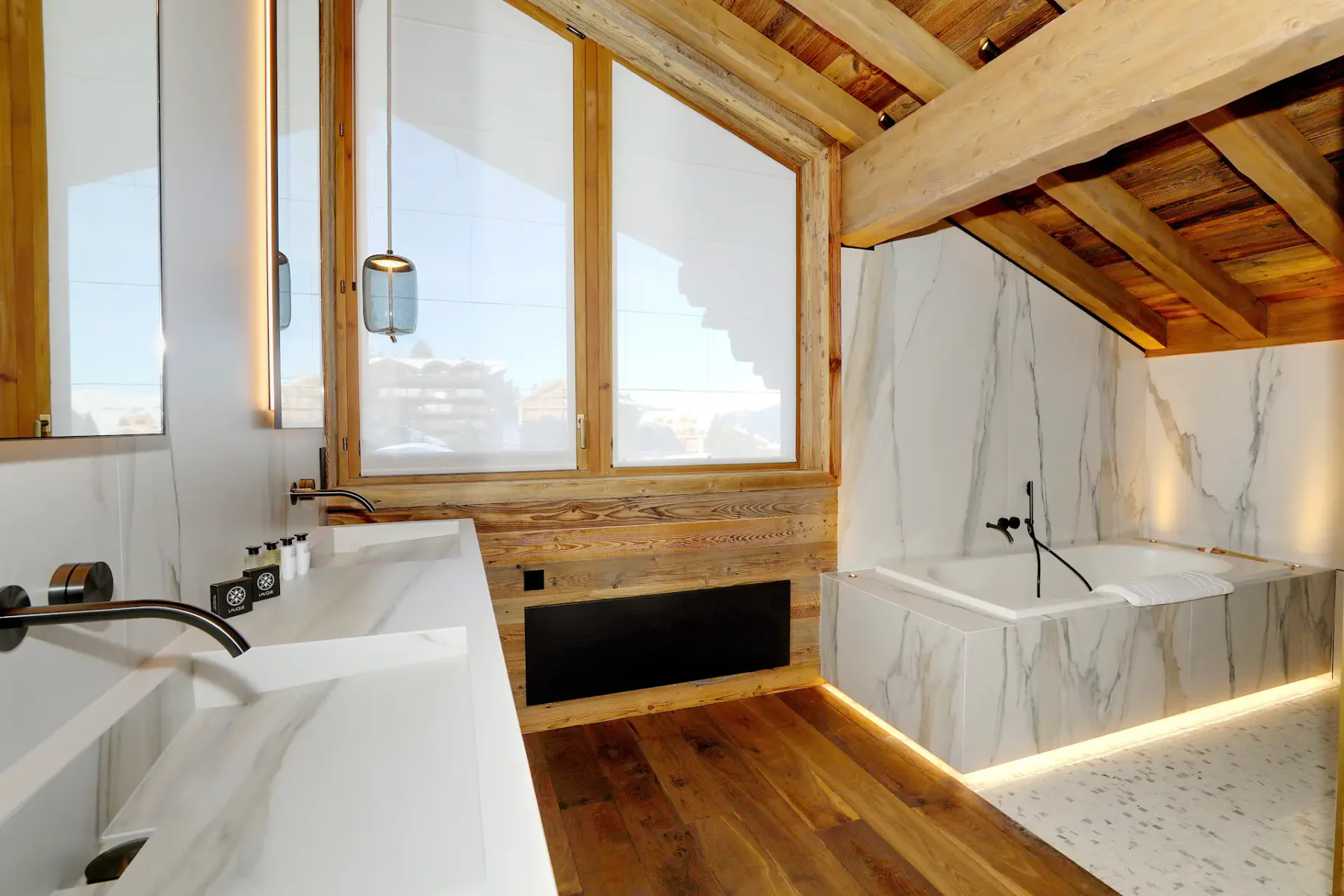 Lujoso baño con vigas de madera, paredes de mármol, dos lavabos y una ventana grande. Cuenta con una bañera moderna y suelo de madera.