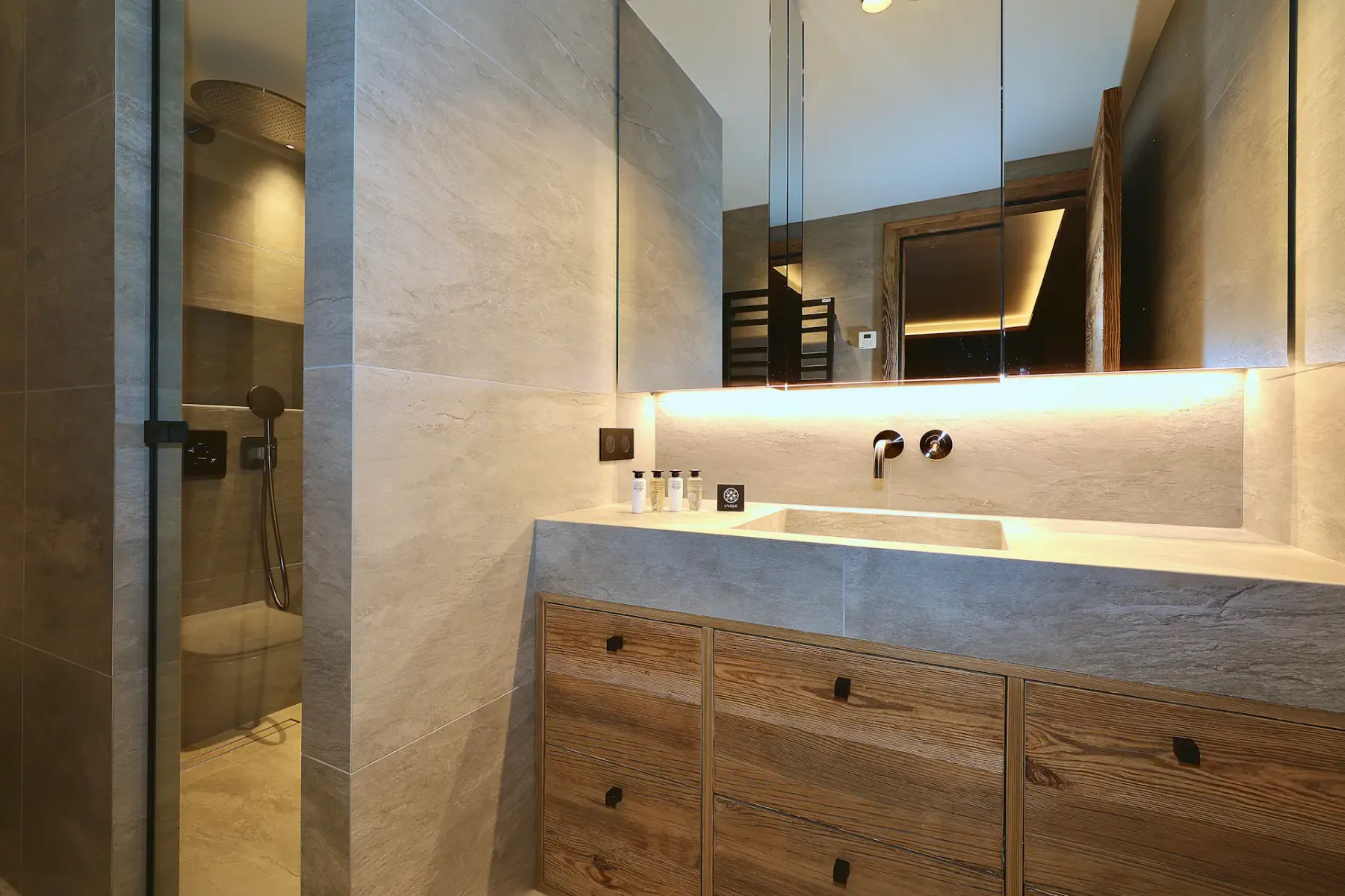 Baño moderno con un mueble de lavabo de madera, un espejo grande y una ducha con cerramiento de cristal. La iluminación suave crea un ambiente cálido.