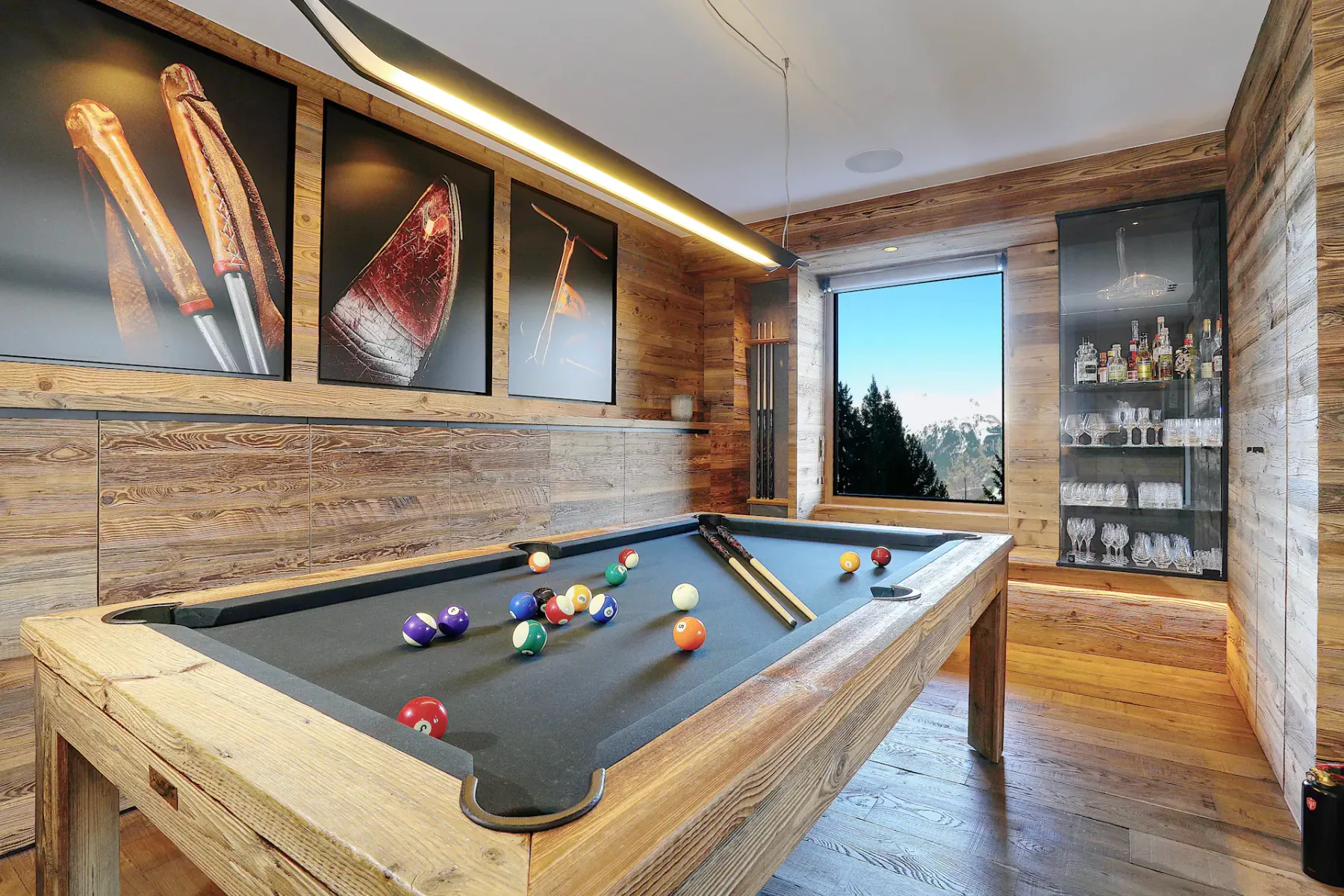 Sala de juegos rústica con una mesa de billar de madera, bolas de colores, tacos y decoración artística en las paredes. Una ventana ofrece vistas a los árboles y las montañas.