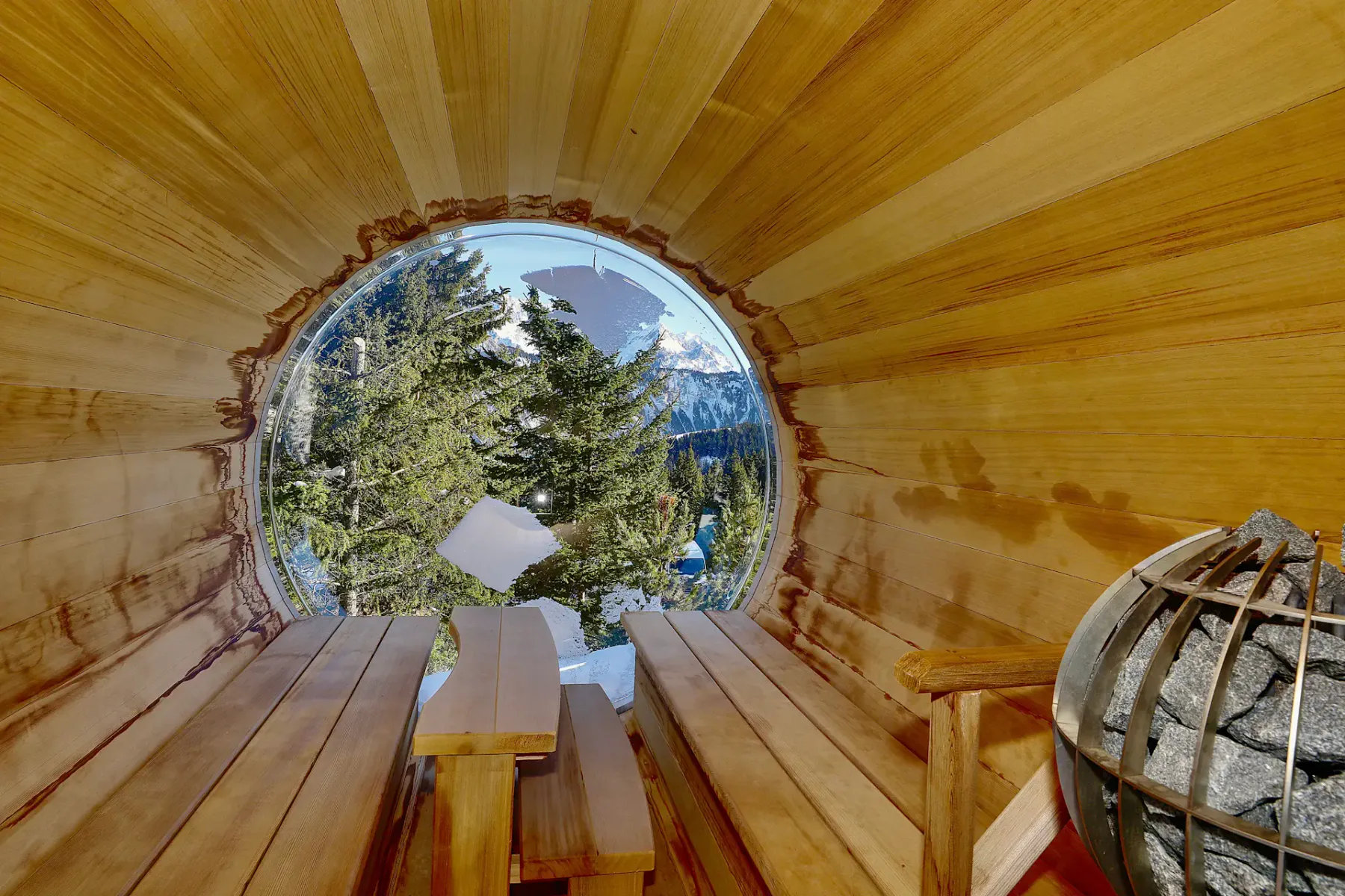 Interior de una sauna de madera en forma de barril, con una ventana con vistas a montañas nevadas y árboles; a la derecha se ven piedras sobre un calentador.