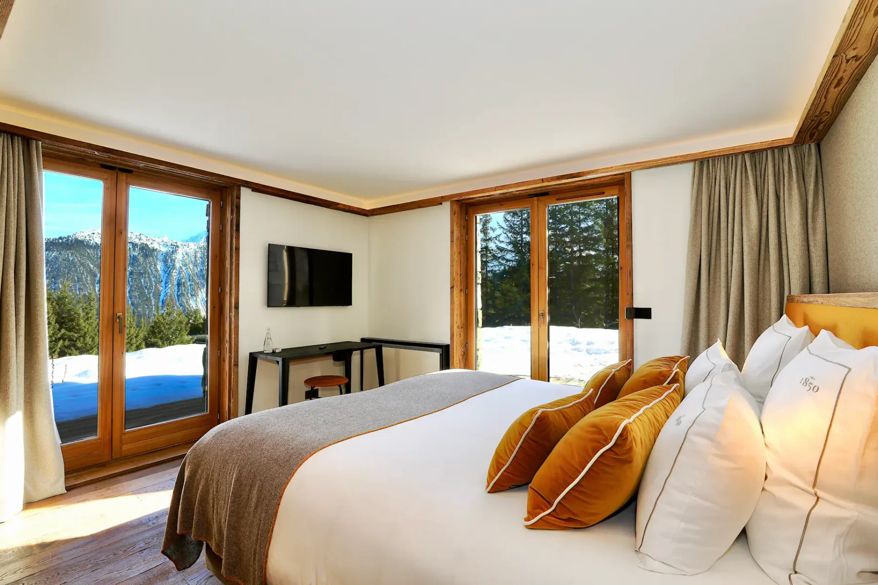 Acogedor dormitorio con una cama grande, almohadas y una manta. Detalles de madera y grandes ventanas con vistas a montañas nevadas y árboles.