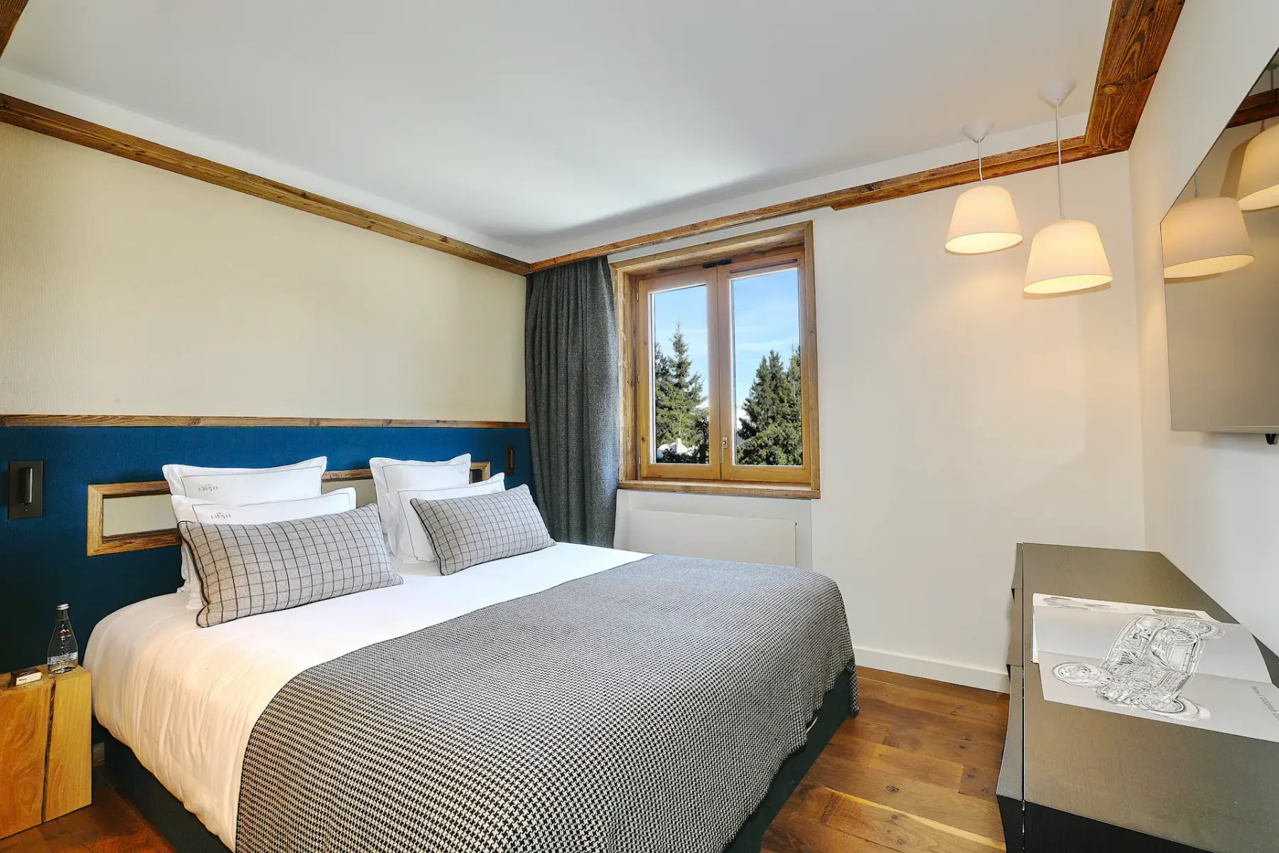 Acogedor dormitorio con una cama doble, almohadas de cuadros, detalles de madera, una ventana con vistas a los árboles y luminarias modernas.
