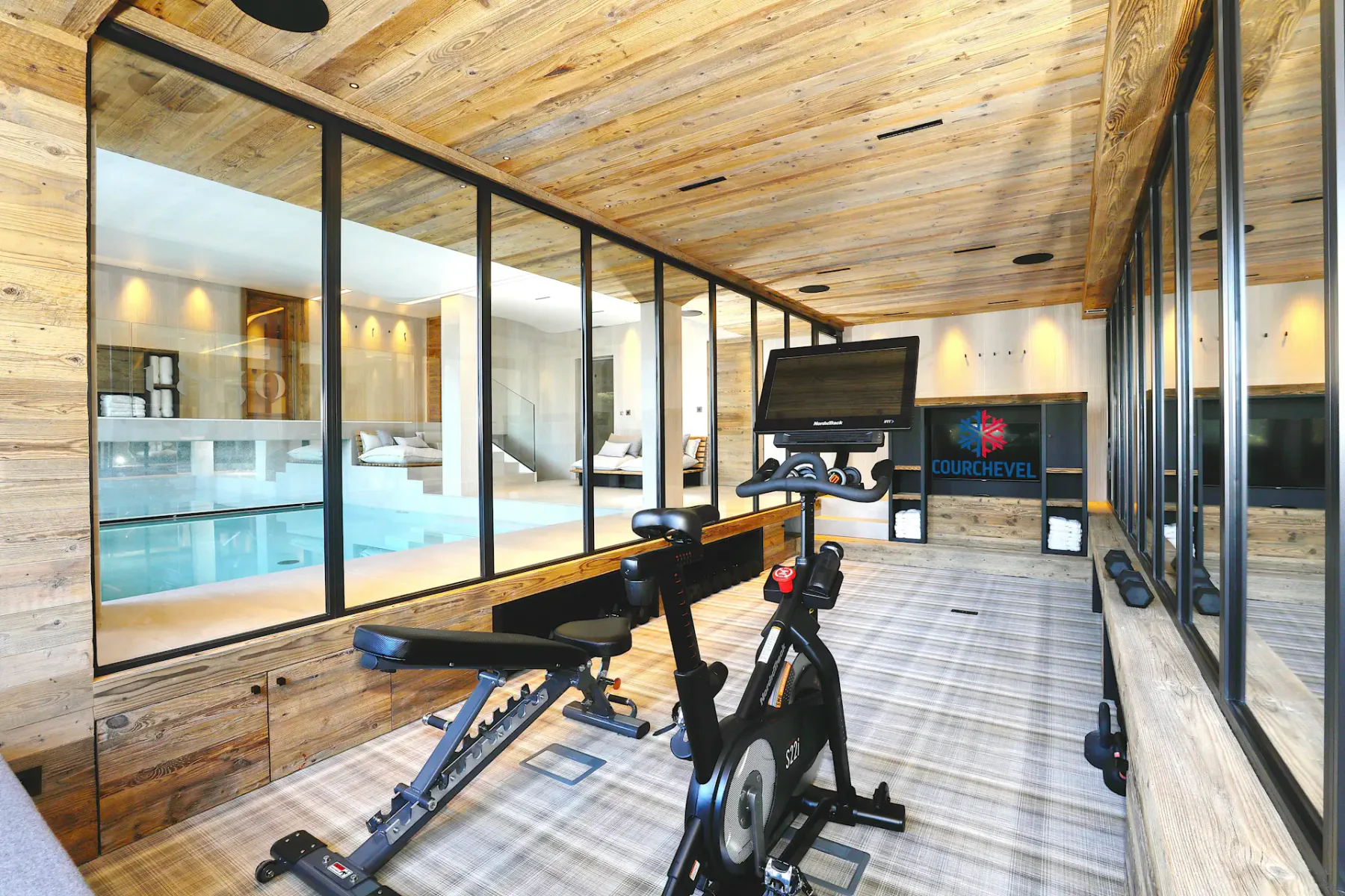 Gimnasio en casa con bicicletas estáticas orientadas hacia un gran ventanal con vistas a una piscina interior. La estancia cuenta con paredes de madera y moqueta de cuadros.