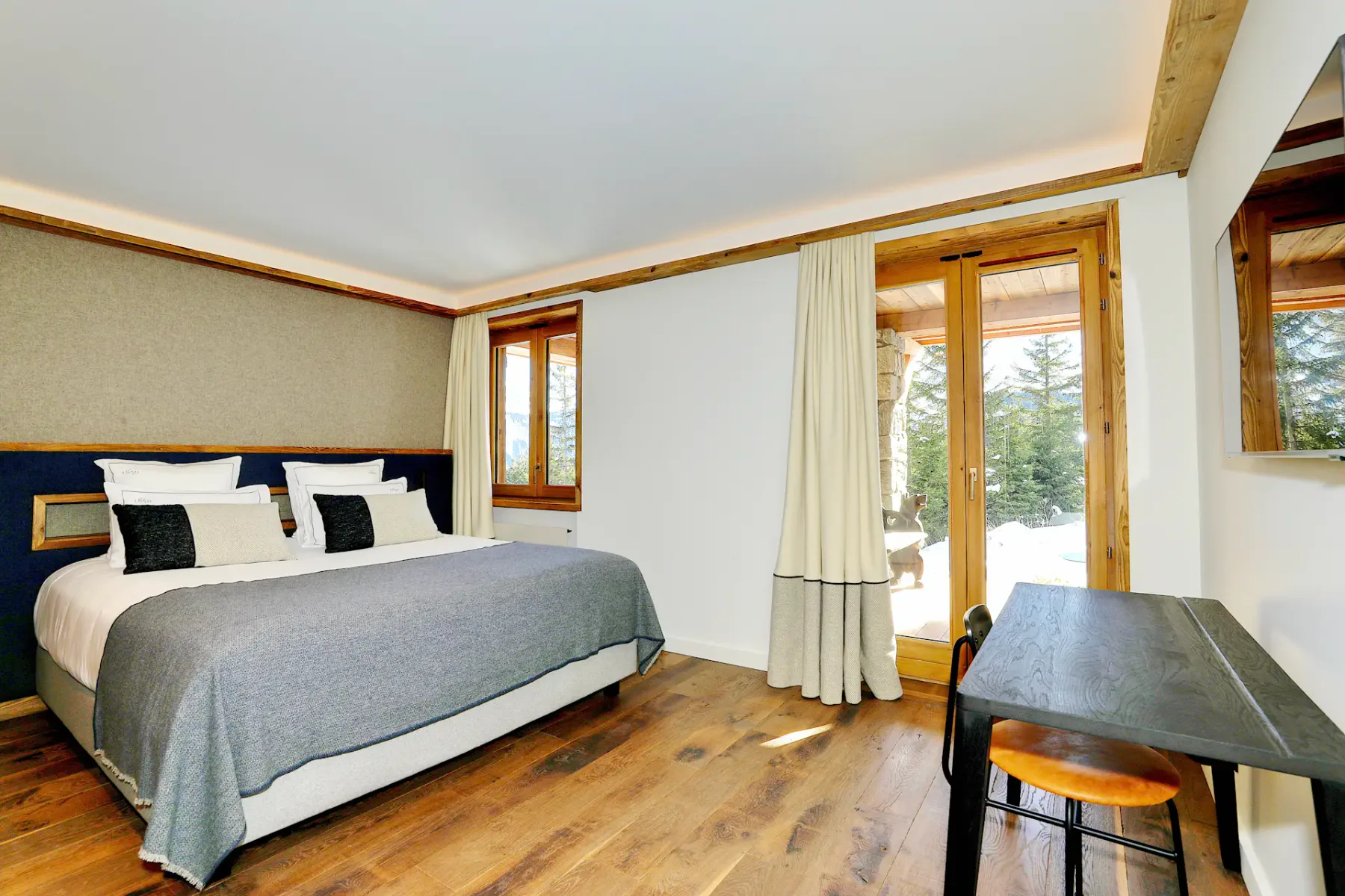 Acogedor dormitorio con una cama grande, suelo de madera, escritorio y puertas de cristal que dan a un exterior nevado. Los tonos neutros y la luz natural llenan el espacio.