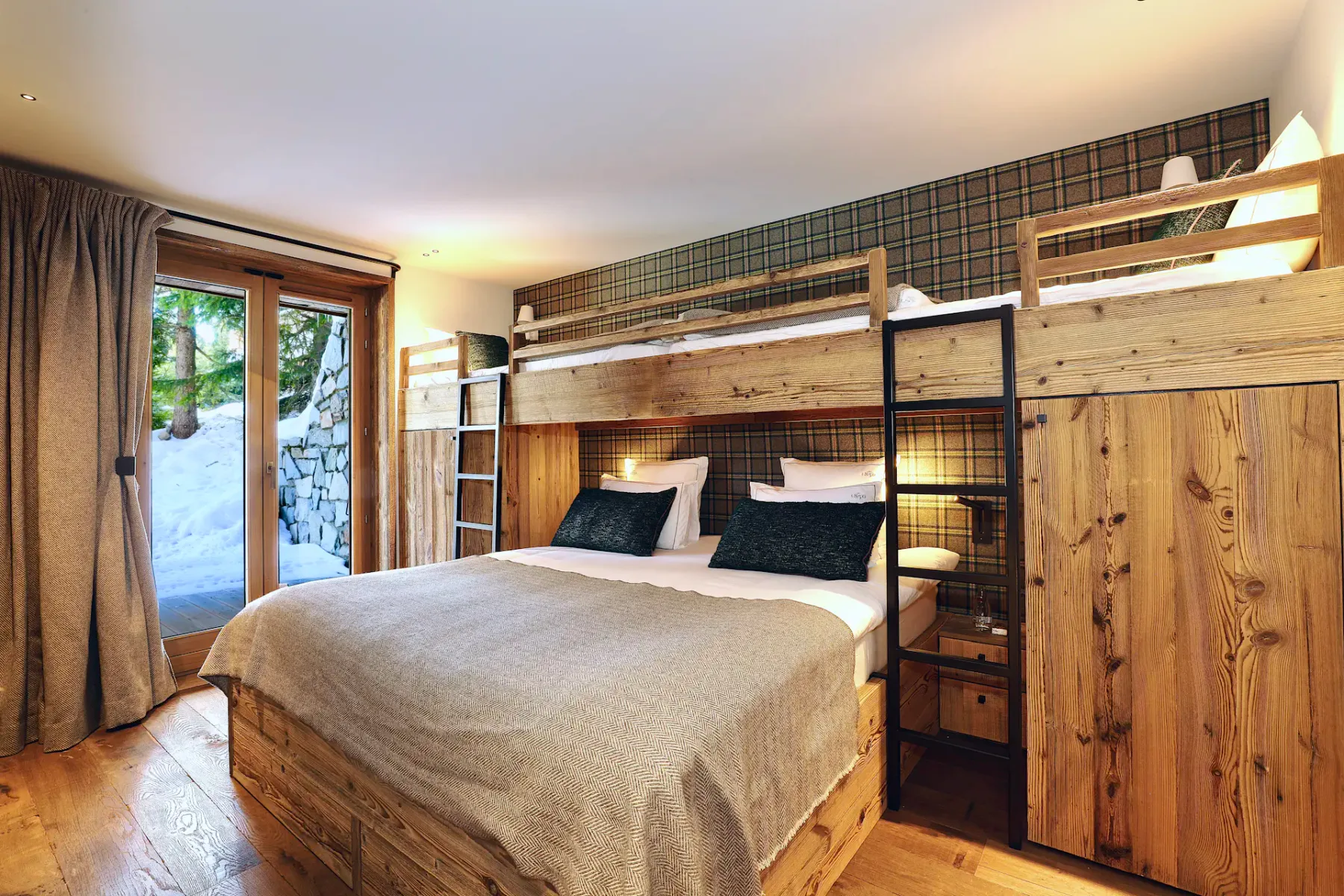 Acogedor dormitorio con literas de madera, una cama doble, detalles de cuadros y vistas a un paisaje nevado a través de puertas de cristal.