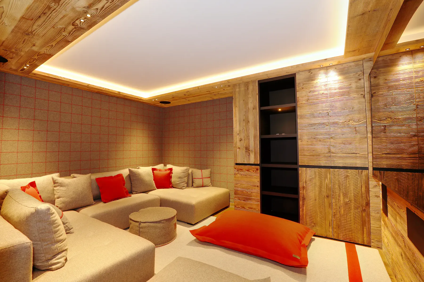 Acogedora estancia con sofá modular beige, cojines rojos, paredes de madera e iluminación suave. Gran cojín de suelo rojo y pequeña mesa redonda.