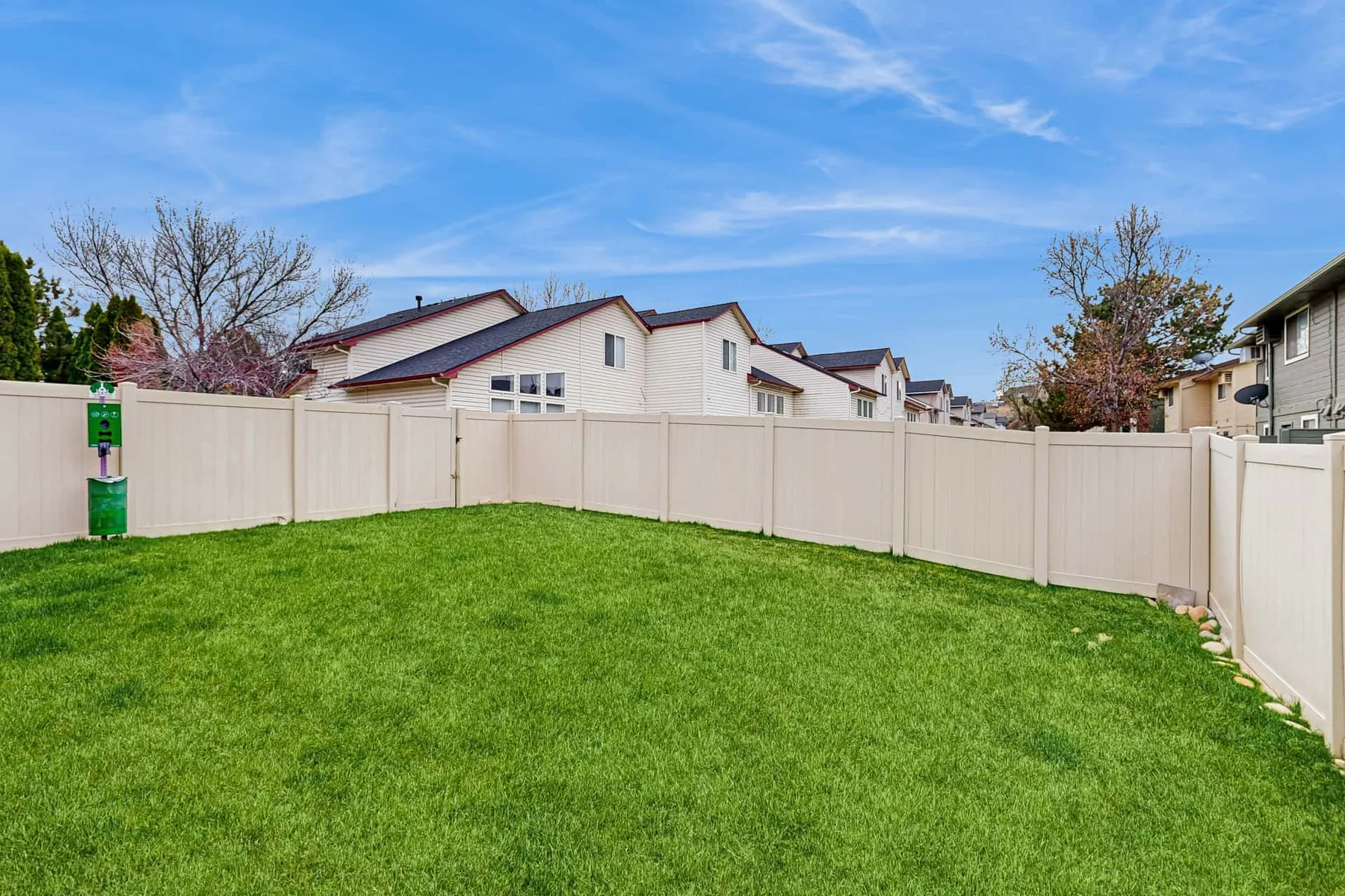 A fenced grassy yard sits beside a row of light-colored homes under a blue sky.