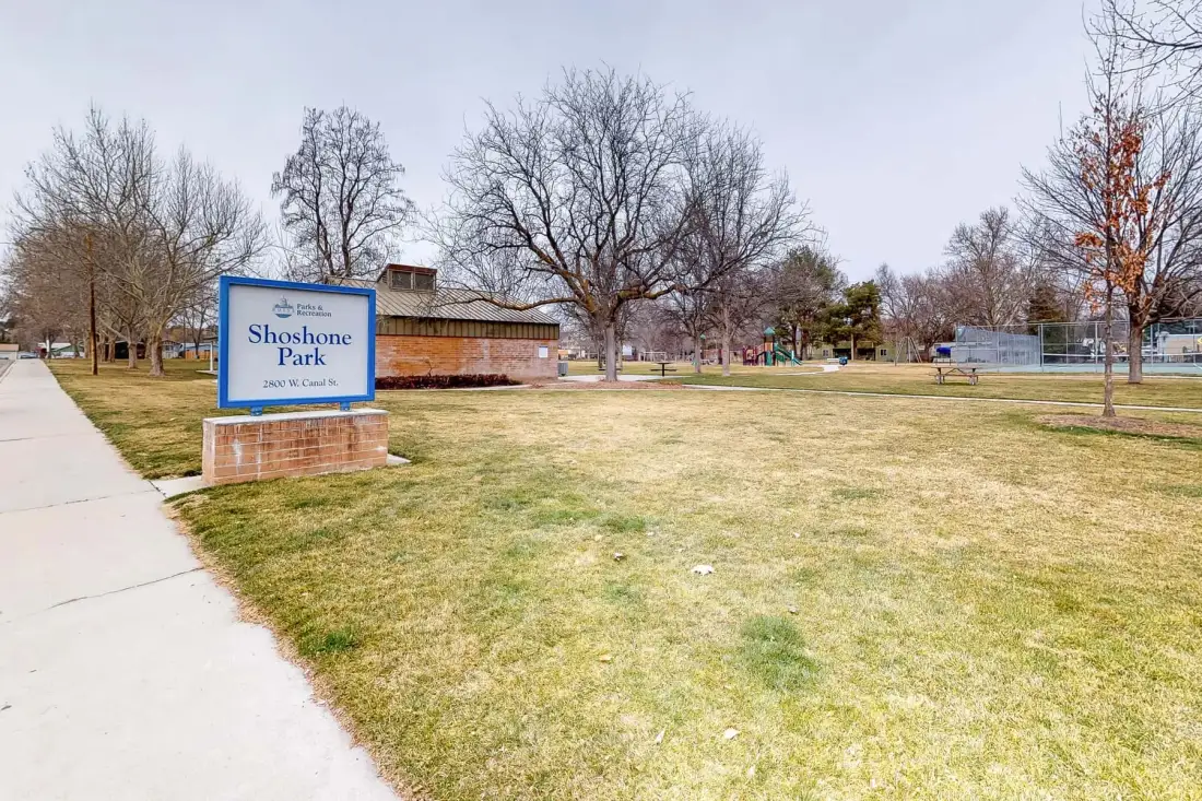 A grassy park with a sidewalk, trees, and a sign at the entrance.