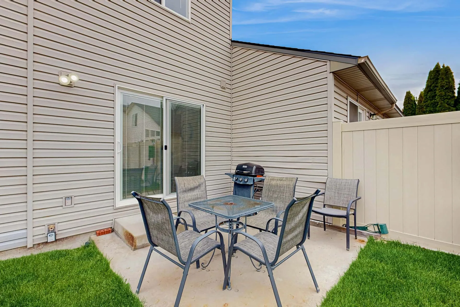 A patio dining area with metal seating sits beside the house and a white fence.